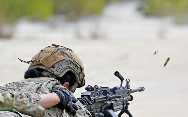 Two military personnel in full combat gear are conducting a live-fire training exercise in a prone position on a sandy range. The shooter in the foreground is actively firing a light machine gun, with spent brass casings visibly ejecting from the weapon. A wooden ammunition crate is positioned to the right side of the firing position.
