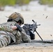 Two military personnel in full combat gear are conducting a live-fire training exercise in a prone position on a sandy range. The shooter in the foreground is actively firing a light machine gun, with spent brass casings visibly ejecting from the weapon. A wooden ammunition crate is positioned to the right side of the firing position.