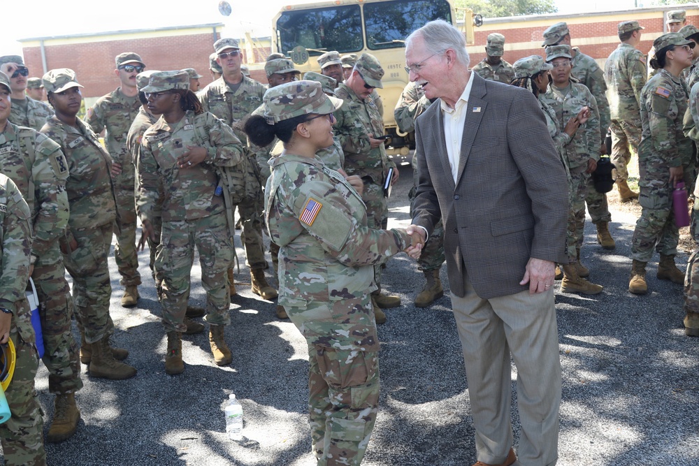 A civilian official in a brown sport coat is shaking hands with a female U.S. Army soldier in MultiCam (OCP) camouflage uniform during what appears to be a formal greeting or send-off event. A large group of uniformed soldiers stands in formation or at ease in the background near a military vehicle. The setting appears to be an outdoor paved area adjacent to a brick building.