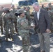 A civilian official in a brown sport coat is shaking hands with a female U.S. Army soldier in MultiCam (OCP) camouflage uniform during what appears to be a formal greeting or send-off event. A large group of uniformed soldiers stands in formation or at ease in the background near a military vehicle. The setting appears to be an outdoor paved area adjacent to a brick building.