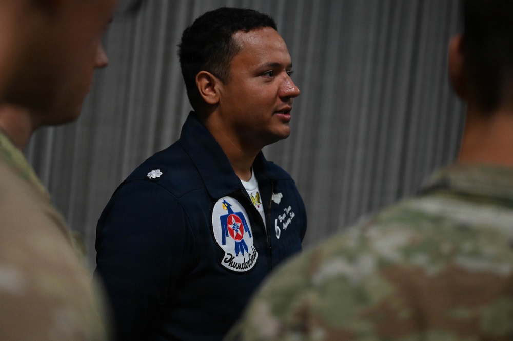 A military aviator wearing a navy blue flight suit with a Thunderbirds patch is speaking to a group of personnel in camouflage uniforms. The individual appears to be a U.S. Air Force Thunderbirds team member, identifiable by the distinctive patch on the left shoulder. The setting appears to be inside a hangar or similar military facility.