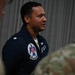 A military aviator wearing a navy blue flight suit with a Thunderbirds patch is speaking to a group of personnel in camouflage uniforms. The individual appears to be a U.S. Air Force Thunderbirds team member, identifiable by the distinctive patch on the left shoulder. The setting appears to be inside a hangar or similar military facility.