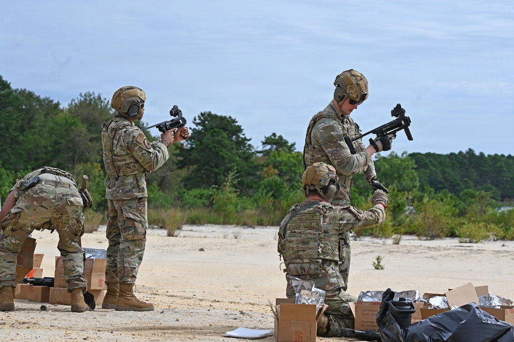 Military personnel in MultiCam/OCP camouflage uniforms and tactical gear are conducting what appears to be a weapons familiarization or qualification exercise at an outdoor range. At least four soldiers are visible, with two standing and handling compact submachine gun-type weapons, one kneeling assisting with equipment, and one bending over supply boxes. The setting is a sandy open area with tree line in the background, consistent with a military training range.