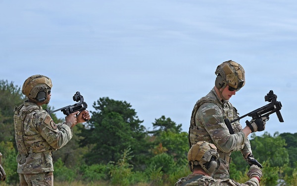 Military personnel in MultiCam/OCP camouflage uniforms and tactical gear are conducting what appears to be a weapons familiarization or qualification exercise at an outdoor range. At least four soldiers are visible, with two standing and handling compact submachine gun-type weapons, one kneeling assisting with equipment, and one bending over supply boxes. The setting is a sandy open area with tree line in the background, consistent with a military training range.