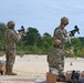 Military personnel in MultiCam/OCP camouflage uniforms and tactical gear are conducting what appears to be a weapons familiarization or qualification exercise at an outdoor range. At least four soldiers are visible, with two standing and handling compact submachine gun-type weapons, one kneeling assisting with equipment, and one bending over supply boxes. The setting is a sandy open area with tree line in the background, consistent with a military training range.
