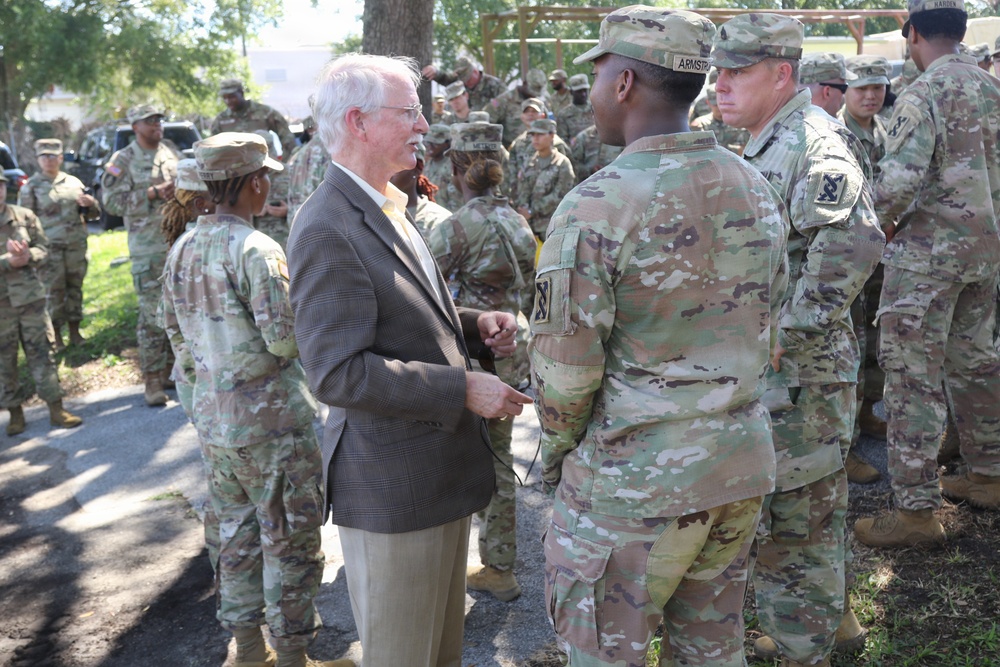 A civilian man in a brown blazer and light-colored trousers is engaged in conversation with U.S. Army soldiers wearing OCP (Operational Camouflage Pattern) uniforms. The gathering appears to be an outdoor military event or briefing, with a large group of soldiers assembled in the background. The soldier facing the civilian has a name tape reading 'ARMSTRONG' and wears a unit patch on the left shoulder.