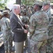A civilian man in a brown blazer and light-colored trousers is engaged in conversation with U.S. Army soldiers wearing OCP (Operational Camouflage Pattern) uniforms. The gathering appears to be an outdoor military event or briefing, with a large group of soldiers assembled in the background. The soldier facing the civilian has a name tape reading 'ARMSTRONG' and wears a unit patch on the left shoulder.