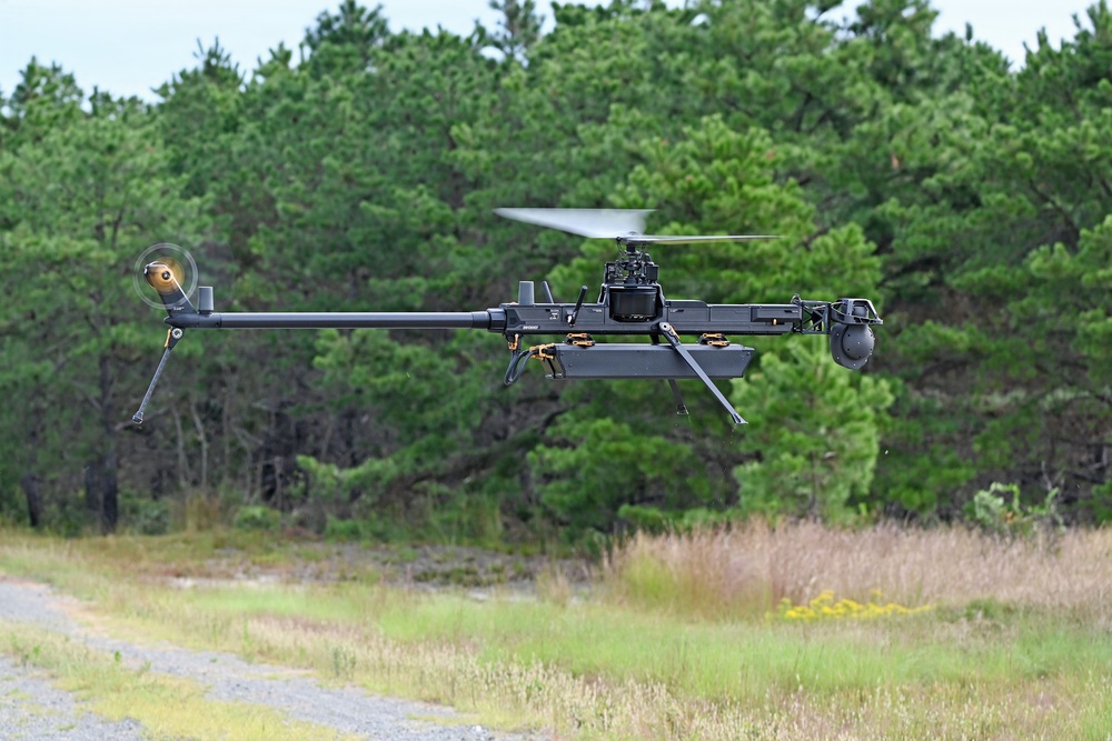 A military or tactical unmanned aerial vehicle (UAV) is captured in flight at low altitude over a grassy clearing bordered by pine trees. The aircraft features a single main rotor configuration with a tail rotor, resembling a helicopter-style drone with a dark matte finish. It appears to be equipped with sensor payloads or camera systems mounted on its frame.