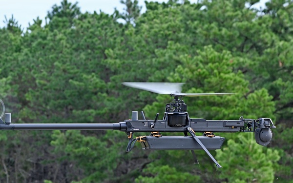 A military or tactical unmanned aerial vehicle (UAV) is captured in flight at low altitude over a grassy clearing bordered by pine trees. The aircraft features a single main rotor configuration with a tail rotor, resembling a helicopter-style drone with a dark matte finish. It appears to be equipped with sensor payloads or camera systems mounted on its frame.