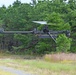A military or tactical unmanned aerial vehicle (UAV) is captured in flight at low altitude over a grassy clearing bordered by pine trees. The aircraft features a single main rotor configuration with a tail rotor, resembling a helicopter-style drone with a dark matte finish. It appears to be equipped with sensor payloads or camera systems mounted on its frame.