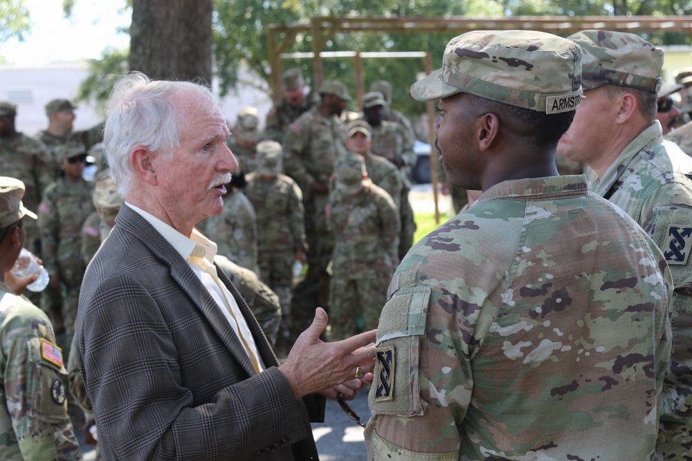 An elderly civilian man in a suit is engaged in conversation with U.S. Army soldiers in Operational Camouflage Pattern (OCP) uniforms outdoors. The central soldier facing away has a name tape reading 'ARMSTR' (Armstrong) and displays a unit patch on the left shoulder. A large group of soldiers is visible in the background, suggesting a formal gathering or event.