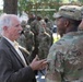 An elderly civilian man in a suit is engaged in conversation with U.S. Army soldiers in Operational Camouflage Pattern (OCP) uniforms outdoors. The central soldier facing away has a name tape reading 'ARMSTR' (Armstrong) and displays a unit patch on the left shoulder. A large group of soldiers is visible in the background, suggesting a formal gathering or event.