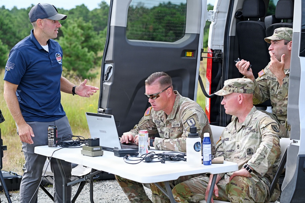 A civilian UAS (Unmanned Aircraft Systems) specialist is briefing three U.S. Army soldiers at an outdoor field operations setup. The group is gathered around a folding table equipped with a laptop and various equipment near a white van. The setting appears to be a training or operational exercise in a wooded area.