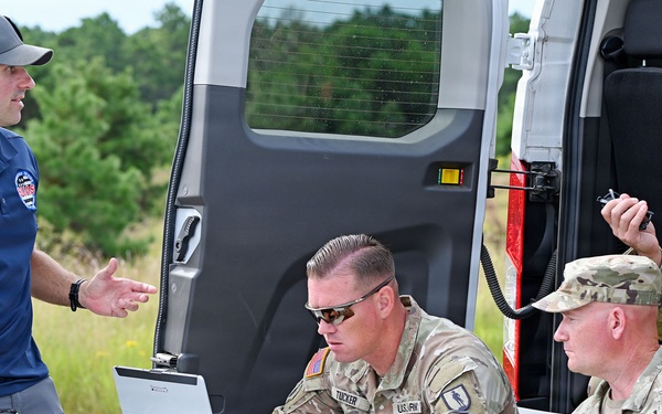 A civilian UAS (Unmanned Aircraft Systems) specialist is briefing three U.S. Army soldiers at an outdoor field operations setup. The group is gathered around a folding table equipped with a laptop and various equipment near a white van. The setting appears to be a training or operational exercise in a wooded area.
