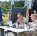 A civilian UAS (Unmanned Aircraft Systems) specialist is briefing three U.S. Army soldiers at an outdoor field operations setup. The group is gathered around a folding table equipped with a laptop and various equipment near a white van. The setting appears to be a training or operational exercise in a wooded area.