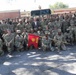 A large group of U.S. Army soldiers in OCP (Operational Camouflage Pattern) uniforms pose for a formal unit photograph. Two civilians in suits are visible among the military personnel, suggesting a dignitary or official visit. A red guidon flag bearing the number '489' and what appears to be an ordnance or engineer branch insignia is prominently displayed in the foreground.