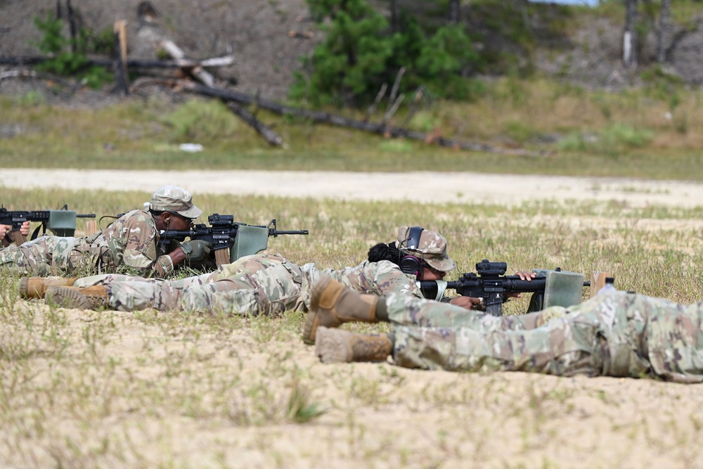 Multiple soldiers in U.S. Army OCP (Operational Camouflage Pattern) uniforms are in the prone firing position on a sandy range, aiming rifles downrange. The scene appears to be a live-fire qualification or training exercise at a military shooting range. At least three soldiers are visible in the foreground, with a possible observer or additional personnel visible in the background.