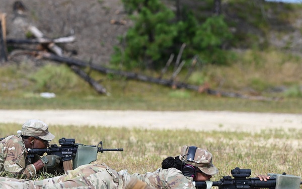 Multiple soldiers in U.S. Army OCP (Operational Camouflage Pattern) uniforms are in the prone firing position on a sandy range, aiming rifles downrange. The scene appears to be a live-fire qualification or training exercise at a military shooting range. At least three soldiers are visible in the foreground, with a possible observer or additional personnel visible in the background.