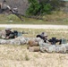 Multiple soldiers in U.S. Army OCP (Operational Camouflage Pattern) uniforms are in the prone firing position on a sandy range, aiming rifles downrange. The scene appears to be a live-fire qualification or training exercise at a military shooting range. At least three soldiers are visible in the foreground, with a possible observer or additional personnel visible in the background.