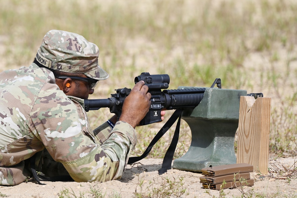 A U.S. Army soldier in MultiCam/OCP camouflage uniform is in a prone firing position at a shooting range, aiming a rifle equipped with an optic sight. The soldier is using a green sandbag rest support and has multiple loaded magazines staged nearby. The name tape on the cap reads 'JOHNSON' and the soldier is wearing ear protection and eye protection.
