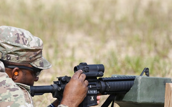 A U.S. Army soldier in MultiCam/OCP camouflage uniform is in a prone firing position at a shooting range, aiming a rifle equipped with an optic sight. The soldier is using a green sandbag rest support and has multiple loaded magazines staged nearby. The name tape on the cap reads 'JOHNSON' and the soldier is wearing ear protection and eye protection.