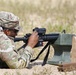 A U.S. Army soldier in MultiCam/OCP camouflage uniform is in a prone firing position at a shooting range, aiming a rifle equipped with an optic sight. The soldier is using a green sandbag rest support and has multiple loaded magazines staged nearby. The name tape on the cap reads 'JOHNSON' and the soldier is wearing ear protection and eye protection.