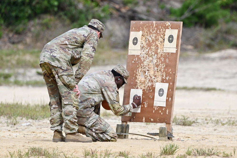 Two military personnel in OCP (Operational Camouflage Pattern) uniforms are examining a shooting target board at what appears to be an outdoor firing range. One soldier is kneeling while the other leans over, both inspecting the lower targets on a heavily damaged wooden target stand. The board shows significant bullet damage with wood splinters and four paper bullseye targets attached.