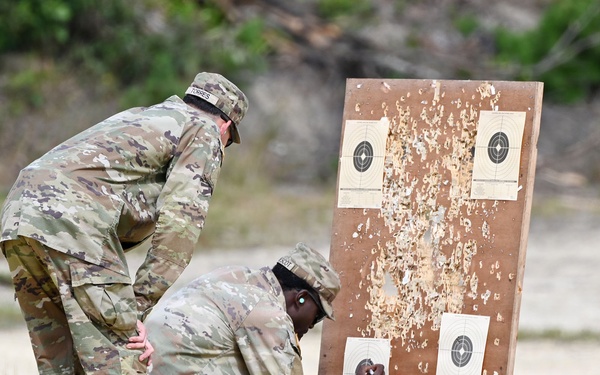 Two military personnel in OCP (Operational Camouflage Pattern) uniforms are examining a shooting target board at what appears to be an outdoor firing range. One soldier is kneeling while the other leans over, both inspecting the lower targets on a heavily damaged wooden target stand. The board shows significant bullet damage with wood splinters and four paper bullseye targets attached.
