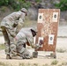 Two military personnel in OCP (Operational Camouflage Pattern) uniforms are examining a shooting target board at what appears to be an outdoor firing range. One soldier is kneeling while the other leans over, both inspecting the lower targets on a heavily damaged wooden target stand. The board shows significant bullet damage with wood splinters and four paper bullseye targets attached.