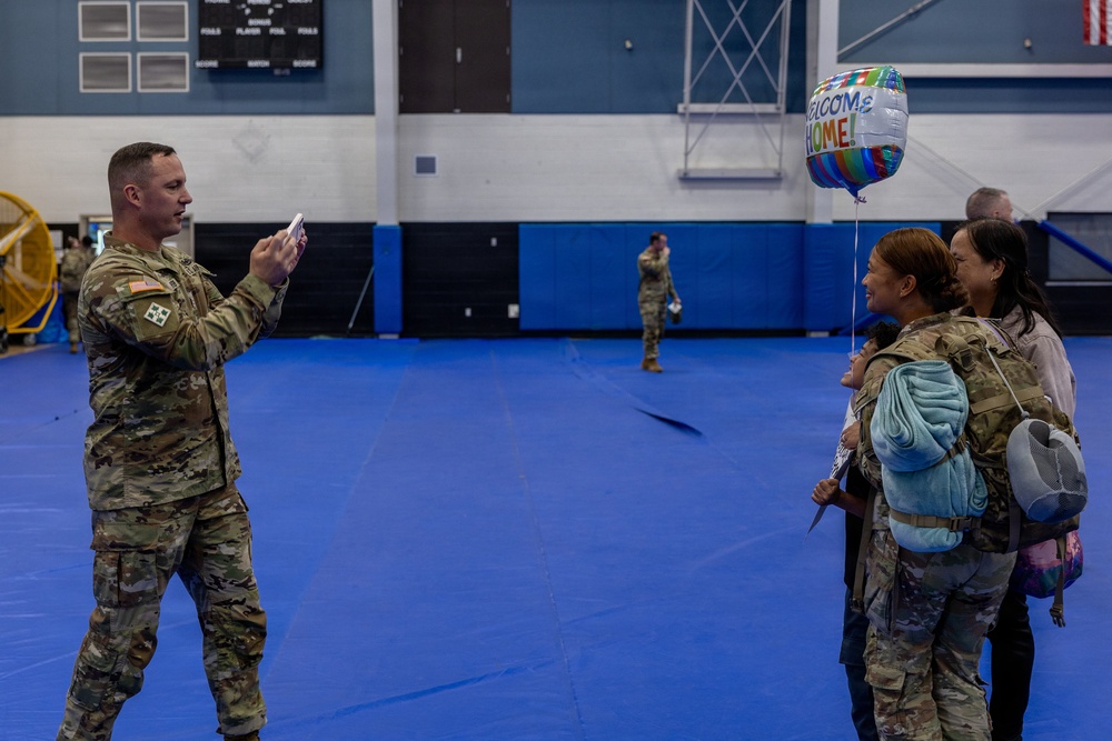 A military homecoming ceremony taking place inside what appears to be a gymnasium or indoor sports facility with blue matted floors. A male soldier in Army Combat Uniform (ACU) is photographing two female soldiers posing together, one of whom is holding a 'Welcome Home' balloon. The scene suggests a unit return from deployment.