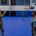 A military homecoming ceremony taking place inside what appears to be a gymnasium or indoor sports facility with blue matted floors. A male soldier in Army Combat Uniform (ACU) is photographing two female soldiers posing together, one of whom is holding a 'Welcome Home' balloon. The scene suggests a unit return from deployment.