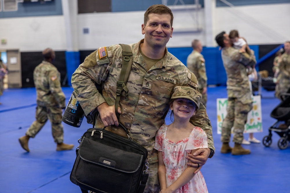 A U.S. Army soldier in OCP (Operational Camouflage Pattern) uniform poses with a young girl wearing his military cap in what appears to be a gymnasium during a homecoming or deployment ceremony. The soldier is smiling and has his arm around the child, who is dressed in a floral dress and also smiling. In the background, other soldiers and civilians can be seen embracing, consistent with a military reunion event.