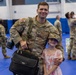 A U.S. Army soldier in OCP (Operational Camouflage Pattern) uniform poses with a young girl wearing his military cap in what appears to be a gymnasium during a homecoming or deployment ceremony. The soldier is smiling and has his arm around the child, who is dressed in a floral dress and also smiling. In the background, other soldiers and civilians can be seen embracing, consistent with a military reunion event.