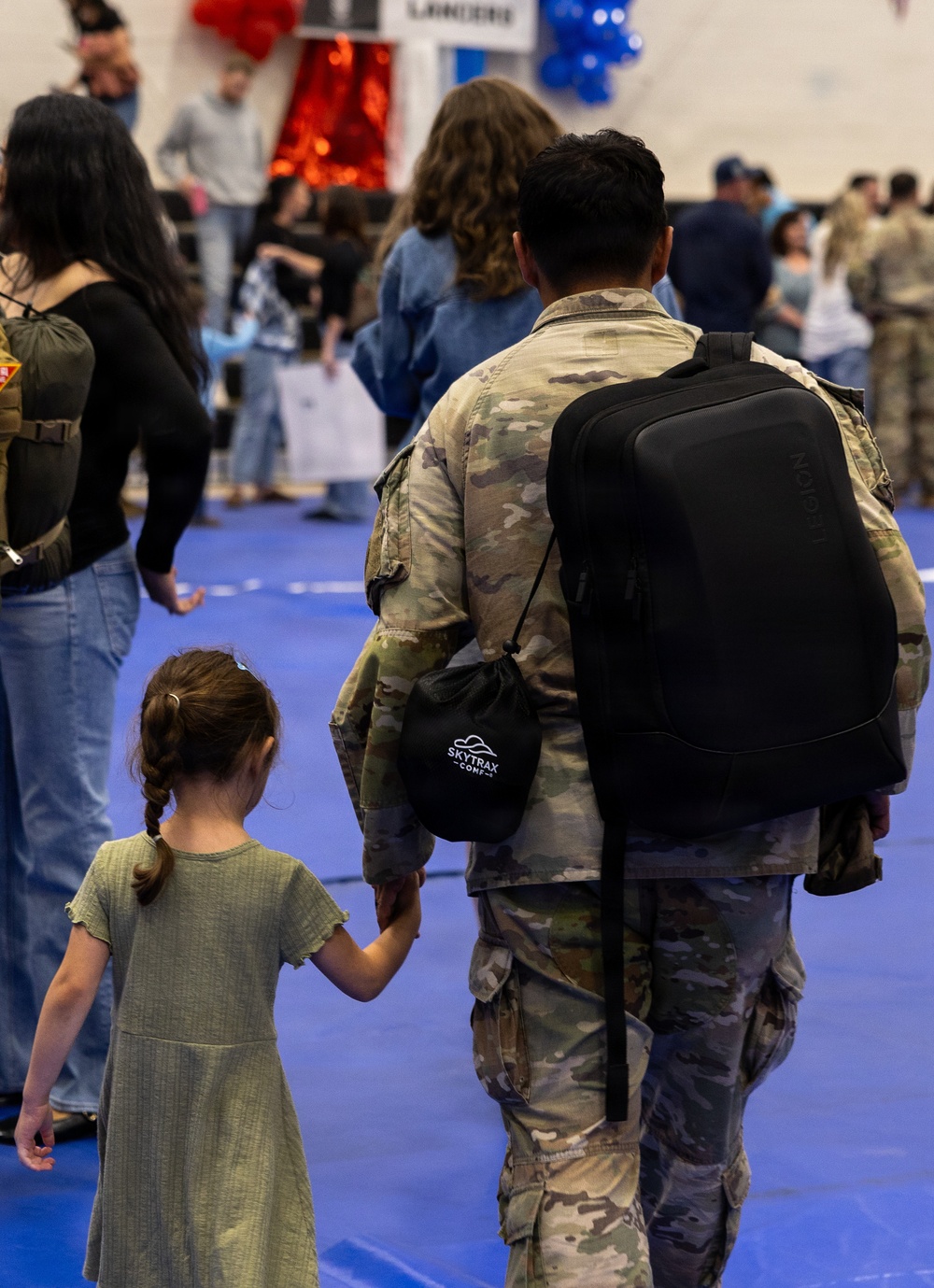 A military service member in OCP (Operational Camouflage Pattern) uniform holds hands with a young girl in a green dress inside what appears to be a gymnasium or indoor event space. The scene suggests a military homecoming or welcome-back ceremony, evidenced by decorative balloons, banners, and a crowd of families in the background. The intimate hand-holding gesture between the soldier and child is the emotional focal point of the image.