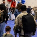A military service member in OCP (Operational Camouflage Pattern) uniform holds hands with a young girl in a green dress inside what appears to be a gymnasium or indoor event space. The scene suggests a military homecoming or welcome-back ceremony, evidenced by decorative balloons, banners, and a crowd of families in the background. The intimate hand-holding gesture between the soldier and child is the emotional focal point of the image.