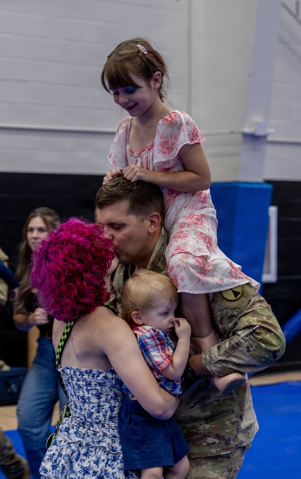 A military service member in camouflage uniform is reuniting with his family in what appears to be a gymnasium or indoor facility. He is kissing a woman with bright pink curly hair while holding a toddler, and a young girl in a floral dress sits on his shoulders. The scene captures an emotional homecoming moment.