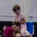 A military service member in camouflage uniform is reuniting with his family in what appears to be a gymnasium or indoor facility. He is kissing a woman with bright pink curly hair while holding a toddler, and a young girl in a floral dress sits on his shoulders. The scene captures an emotional homecoming moment.