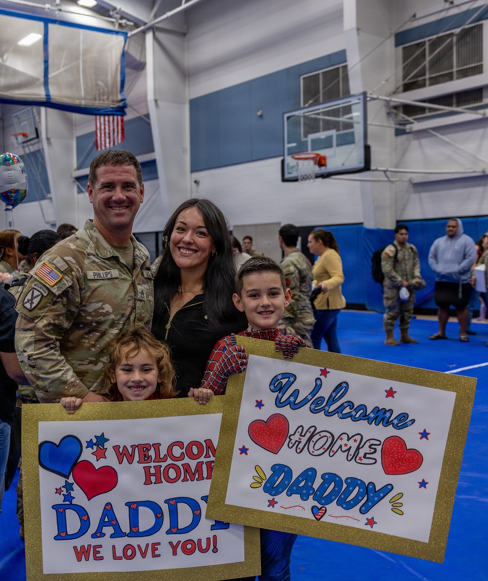 A U.S. Army soldier named Phillips, wearing a 10th Mountain Division patch, is reunited with his family at a military homecoming ceremony held in a gymnasium. His wife and two young children hold handmade signs reading 'Welcome Home Daddy' and 'Welcome Home Daddy We Love You!' decorated with hearts and stars. The background shows other soldiers and families participating in the same welcome home event.