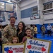 A U.S. Army soldier named Phillips, wearing a 10th Mountain Division patch, is reunited with his family at a military homecoming ceremony held in a gymnasium. His wife and two young children hold handmade signs reading 'Welcome Home Daddy' and 'Welcome Home Daddy We Love You!' decorated with hearts and stars. The background shows other soldiers and families participating in the same welcome home event.