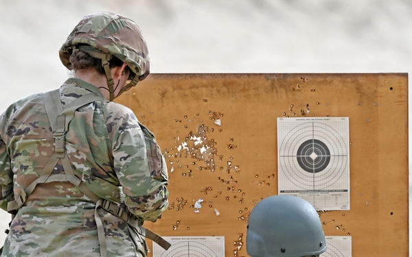 Two military personnel in camouflage uniforms are examining a shooting target board at a firing range. The wooden target board displays multiple paper bullseye targets with numerous bullet holes scattered across the board and targets. One soldier is standing while the other is crouching and pointing at the lower target, apparently assessing shot placement.