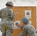 Two military personnel in camouflage uniforms are examining a shooting target board at a firing range. The wooden target board displays multiple paper bullseye targets with numerous bullet holes scattered across the board and targets. One soldier is standing while the other is crouching and pointing at the lower target, apparently assessing shot placement.