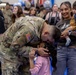 A U.S. Army soldier in OCP (Operational Camouflage Pattern) uniform bends down to kiss a young girl on the head in what appears to be a military homecoming or welcome ceremony. A smiling woman holding a baby stands nearby, watching the emotional reunion. The indoor venue is filled with other people, balloons, and celebratory decorations in the background.