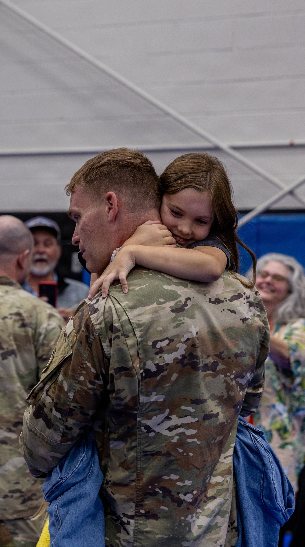 A soldier in OCP (Operational Camouflage Pattern) uniform is holding and embracing a young girl who has her arms wrapped tightly around his neck and is smiling. The scene appears to be a military homecoming or deployment ceremony taking place inside a large indoor venue. Several other people are visible in the background, also appearing joyful and emotional.