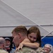 A soldier in OCP (Operational Camouflage Pattern) uniform is holding and embracing a young girl who has her arms wrapped tightly around his neck and is smiling. The scene appears to be a military homecoming or deployment ceremony taking place inside a large indoor venue. Several other people are visible in the background, also appearing joyful and emotional.