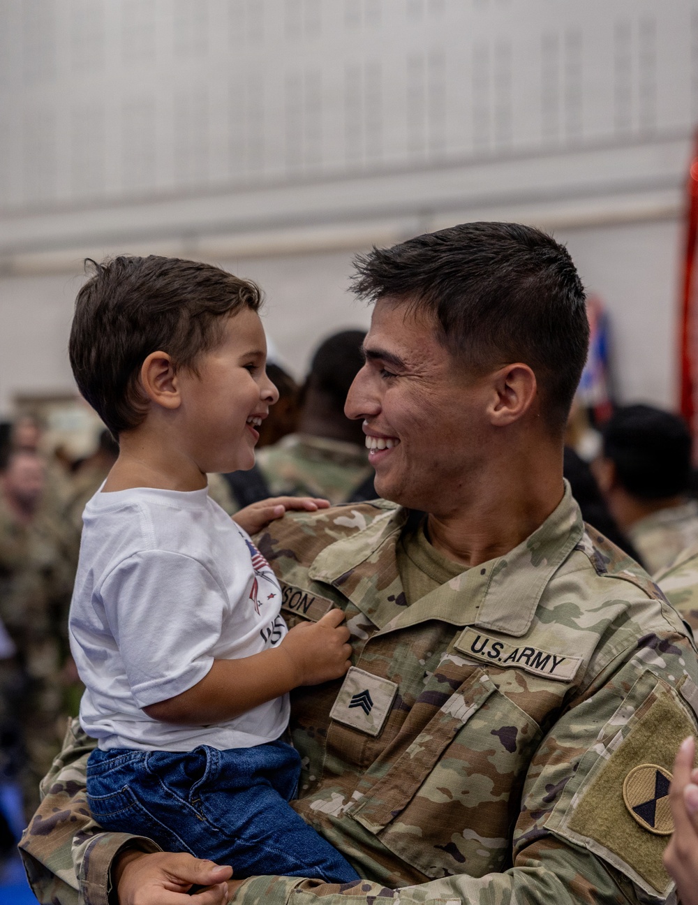 A U.S. Army Sergeant in OCP (Operational Camouflage Pattern) uniform holds a young toddler boy, both smiling and laughing at each other in what appears to be a military reunion or homecoming ceremony. The emotional moment captures a joyful interaction between the soldier and the child, likely a family member. The setting appears to be an indoor military facility with other soldiers visible in the background.