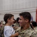 A U.S. Army Sergeant in OCP (Operational Camouflage Pattern) uniform holds a young toddler boy, both smiling and laughing at each other in what appears to be a military reunion or homecoming ceremony. The emotional moment captures a joyful interaction between the soldier and the child, likely a family member. The setting appears to be an indoor military facility with other soldiers visible in the background.