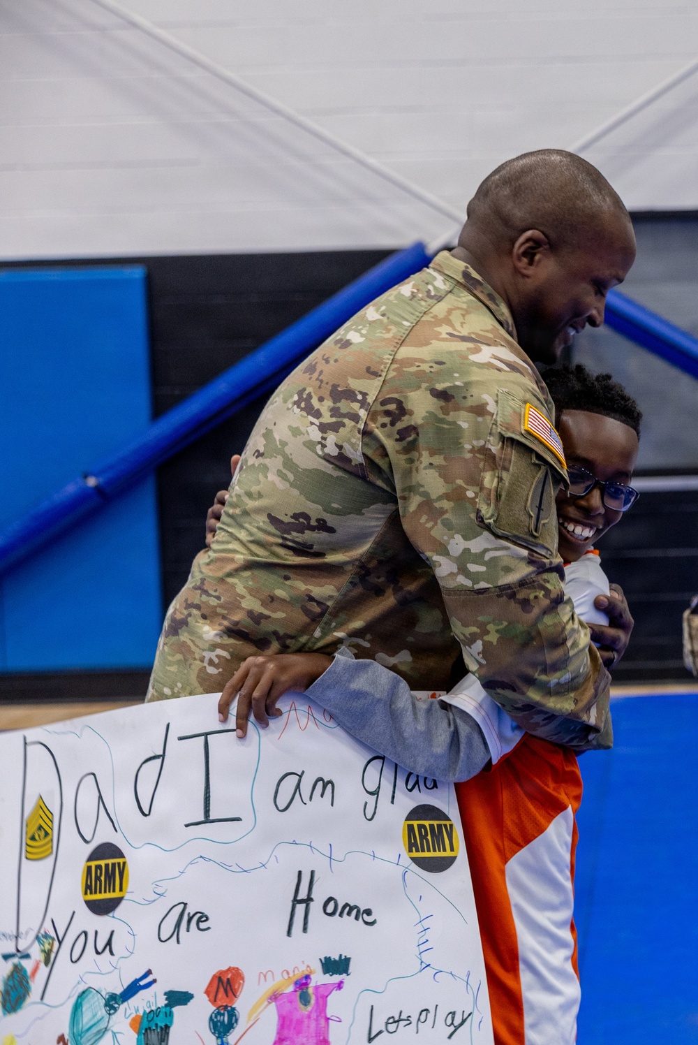 A U.S. Army soldier in OCP (Operational Camouflage Pattern) uniform is embracing a child in what appears to be a military homecoming reunion. The child is holding a handmade sign reading 'Dad I am glad you are Home' decorated with Army stickers and colorful drawings. Both individuals are smiling broadly, capturing an emotional reunion moment likely in a gymnasium or indoor facility.