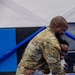 A U.S. Army soldier in OCP (Operational Camouflage Pattern) uniform is embracing a child in what appears to be a military homecoming reunion. The child is holding a handmade sign reading 'Dad I am glad you are Home' decorated with Army stickers and colorful drawings. Both individuals are smiling broadly, capturing an emotional reunion moment likely in a gymnasium or indoor facility.