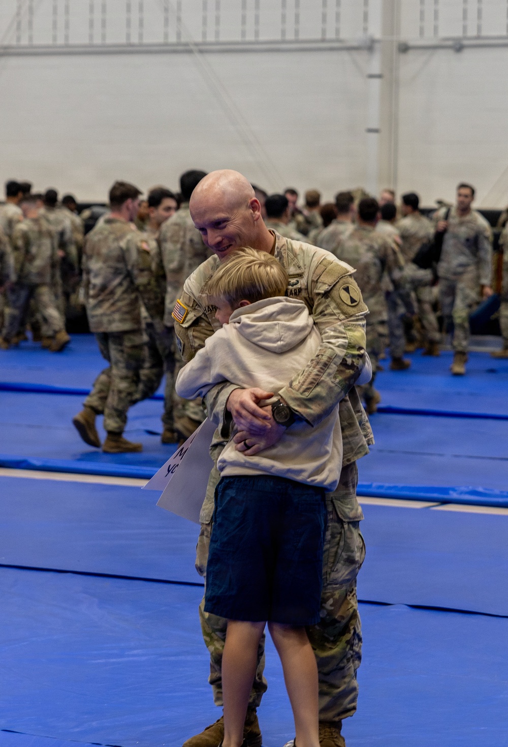 A U.S. Army soldier in OCP (Operational Camouflage Pattern) uniform embraces a young child, likely during a homecoming or deployment ceremony held inside a large indoor facility. The soldier is smiling warmly while hugging the child, who is holding a partially visible sign. Numerous other soldiers in uniform are visible in the background.