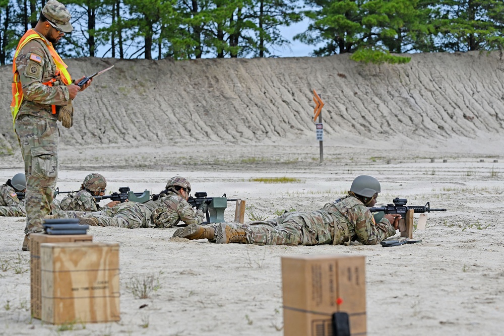 Military personnel are conducting a rifle qualification or marksmanship training exercise on a sandy firing range. Several soldiers are in the prone position aiming rifles downrange while a range safety officer or instructor wearing a high-visibility orange vest observes and records data on a tablet or clipboard. The setting appears to be a standard military live-fire range with a sand berm backstop.